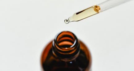 Close-up of a dropper releasing liquid into a glass bottle on a white surface.